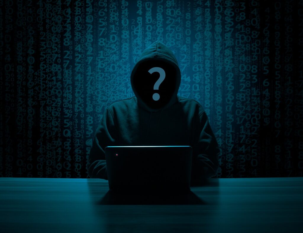 A hooded figure, sitting in the dark in front of a screen with a question-mark over their face.