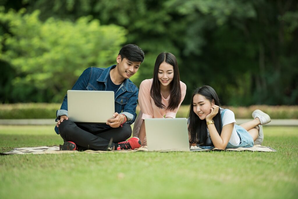 Students sitting on the grass, looking at their laptop screens, smiling.